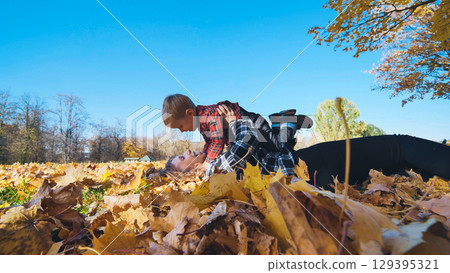 Mother and son enjoying playful moments in a park, lying together on vibrant autumn leaves under a warm, sunny sky 129395321