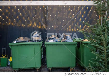 Green garbage bins with trash bags near Christmas tree and festive lights 129397122