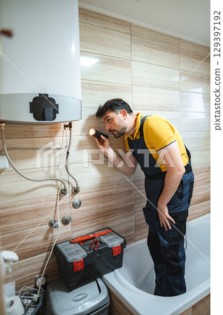 Plumber inspecting water heater in bathroom using flashlight Plumber inspecting water heater in bathroom using flashlight 129397192