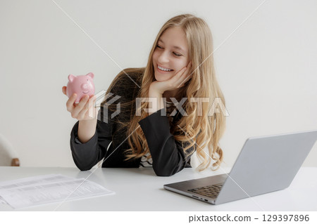 Young woman smiles while holding a piggy bank at her desk with a laptop and financial documents in a bright workspace Young woman smiles while holding a piggy bank at her desk with a laptop and financial documents in a bright workspace 129397896