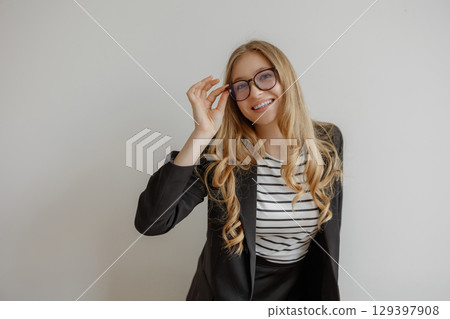 Confident woman in business attire smiling while adjusting her glasses indoors 129397908