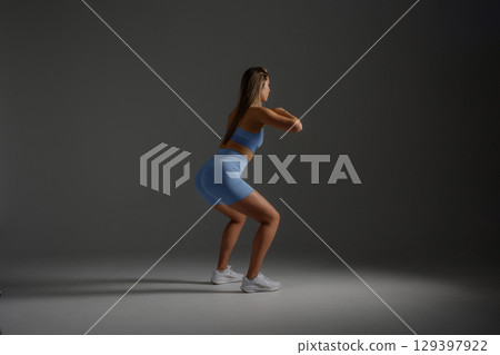Model performs squats during indoor workout session in blue fitness attire at a neutral backdrop 129397922