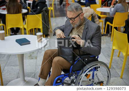 A stylish elderly man in a wheelchair thoughtfully organizes his bag in a busy cafe alone 129398073
