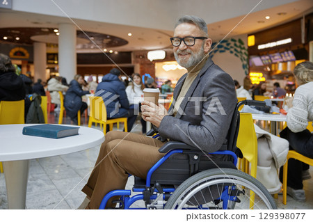 A stylish elderly man in a wheelchair happily enjoying a cup of coffee at a modern cafe 129398077