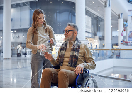 A compassionate woman providing assistance to a senior man who uses a wheelchair at a shopping mall A compassionate woman providing assistance to a senior man who uses a wheelchair at a shopping mall 129398086