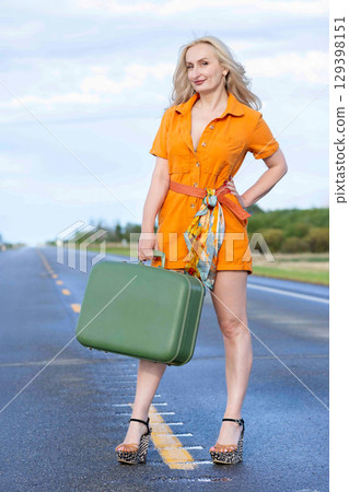 Blonde woman with a green retro suitcase standing on the highway. Blonde woman with a green retro suitcase standing on the highway. 129398151