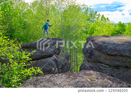 Boy is jumping over big Rocks formation in the green forest. Boy is jumping over big Rocks formation in the green forest. 129398205