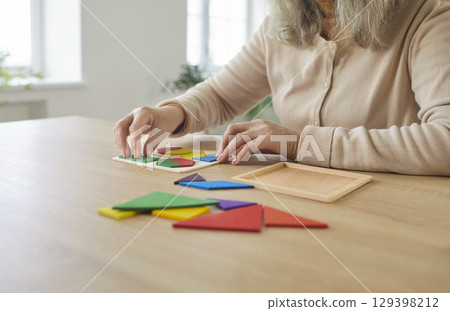 Senior woman sitting playing at table close up, arranging colorful puzzle pieces, working on tangram 129398212