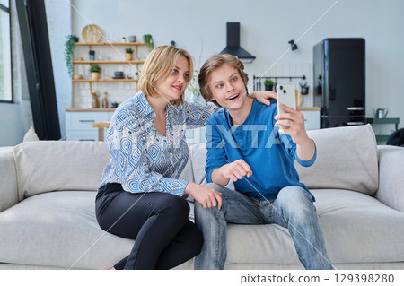 Middle aged mother and teenage son sitting together at home taking selfie photo on smartphone 129398280