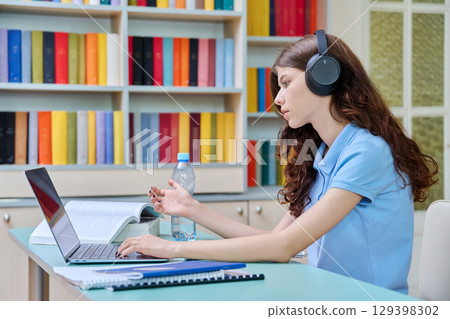 Teenage girl high school student in headphones having video lesson, at library Teenage girl high school student in headphones having video lesson, at library 129398302