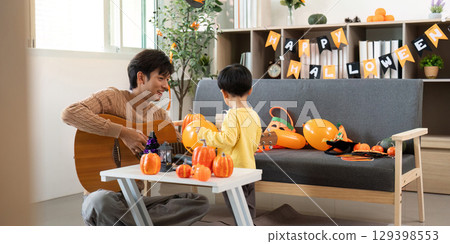 Musical Halloween. Father playing guitar while son enjoys pumpkin decorating. Musical Halloween. Father playing guitar while son enjoys pumpkin decorating. 129398553