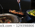 Red king prawns sizzle on a hot grill beside thick beef steaks. A cook uses metal tongs to flip the meat on the grate. Juicy seafood and beef grilling over open fire a delicious mix of surf and turf. 129398866