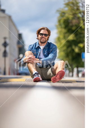 A young man in casual shorts and a denim jacket sits on a pedestrian crossing A young man in casual shorts and a denim jacket sits on a pedestrian crossing 129399752