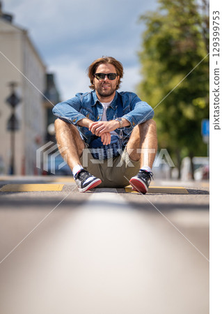 A young man in casual shorts and a denim jacket sits on a pedestrian crossing A young man in casual shorts and a denim jacket sits on a pedestrian crossing 129399753
