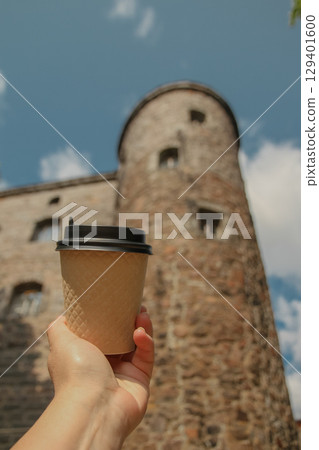 Brown paper cup of coffee in women's hand on against backdrop of old Olaf's Tower in city Vyborg in blur, Russia on sunny day Brown paper cup of coffee in women's hand on against backdrop of old Olaf's Tower in city Vyborg in blur, Russia on sunny day 129401600