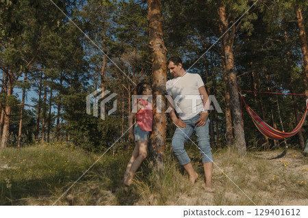 father and daughter stand in a forest clearing near a tree next to a hammock and look at each other in summer, lifestyle 129401612