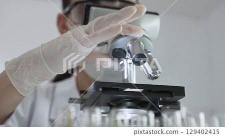 Female scientist wearing white protective gear analyzing test samples under microscope in contemporary research environment, close up of white gloved hand. Medicine and science concept 129402415