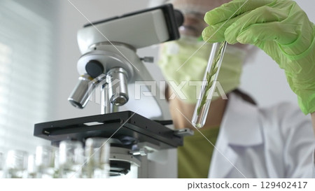 Female scientist in green protective mask and gloves holding test tube with plant sample, researching botany and medical applications using a microscope. Medicine, health care and science concept Female scientist in green protective mask and gloves holding test tube with plant sample, researching botany and medical applications using a microscope. Medicine, health care and science concept 129402417