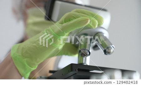 Close up of green gloved hands of a scientist adjusting a microscope in a laboratory, emphasizing the significance of scientific research and analysis. Medicine, healthcare and science concept 129402418