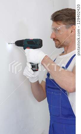 Professional male construction worker in a blue jumpsuit, safety goggles, and work gloves is screwing a screw into the wall using a cordless screwdriver. Home renovation concept Professional male construction worker in a blue jumpsuit, safety goggles, and work gloves is screwing a screw into the wall using a cordless screwdriver. Home renovation concept 129402420