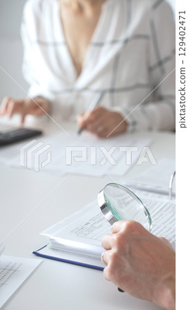 Professional accountants are working together, using a magnifying glass and a calculator, to audit financial reports in office, close up vertical view. Audit and taxes concept 129402471
