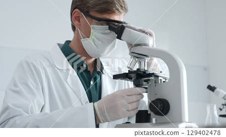 Adult male scientist wearing white mask, lab coat and protective glasses using microscope, doing scientific research in modern laboratory. Medicine, health care and science concept Adult male scientist wearing white mask, lab coat and protective glasses using microscope, doing scientific research in modern laboratory. Medicine, health care and science concept 129402478