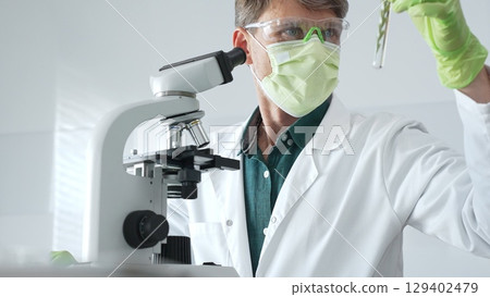 Masked senior man researcher examining green botanical samples in test tube in laboratory during biological analysis. Medicine, health care and science concept 129402479