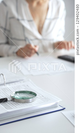 Magnifying glass on financial documents, with an accountant using a calculator and taking notes, emphasizing accounting, finance, and tax preparation, close up vertical view. Audit and taxes Magnifying glass on financial documents, with an accountant using a calculator and taking notes, emphasizing accounting, finance, and tax preparation, close up vertical view. Audit and taxes 129402480