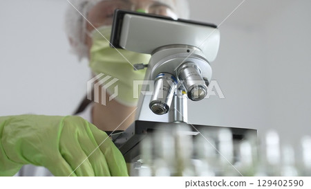 Female scientist wearing green mask and protective glasses using microscope in laboratory, doing scientific research. Medicine, healthcare and science concept 129402590