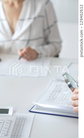 Professional accountants are working together, using a magnifying glass and a calculator, to audit financial reports in office, close up vertical view. Audit and taxes concept Professional accountants are working together, using a magnifying glass and a calculator, to audit financial reports in office, close up vertical view. Audit and taxes concept 129402612