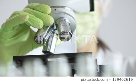 Close up of green gloved hands of a scientist adjusting a microscope in a laboratory, emphasizing the significance of scientific research and analysis. Medicine, healthcare and science concept Close up of green gloved hands of a scientist adjusting a microscope in a laboratory, emphasizing the significance of scientific research and analysis. Medicine, healthcare and science concept 129402619