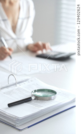 Close up of a magnifying glass on financial documents, with an accountant using a calculator and taking notes, emphasizing accounting, finance, and tax preparation. Audit and taxes in business 129402624