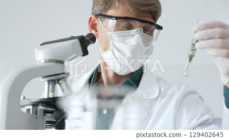 Adult male scientist in mask and gloves holding test tube with green plant sample, conducting laboratory research with a microscope. Medicine, health care and science concept 129402645