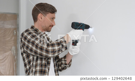 Male construction worker in checked shirt and safety gloves is screwing a screw into the wall using a cordless screwdriver, carrying out a careful home renovation. Portrait view Male construction worker in checked shirt and safety gloves is screwing a screw into the wall using a cordless screwdriver, carrying out a careful home renovation. Portrait view 129402648