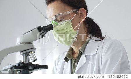 Female professional scientist examining specimen, placing slide under microscope while wearing green protective gear in laboratory workspace. Medicine, health care and science concept 129402652