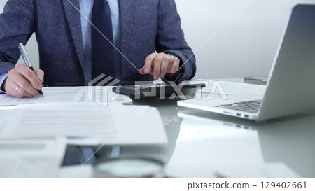 Businessman or accountant wearing blue suit is working with laptop, calculator and documents in office. Low lighting, close-up view. Business people, audit concept Businessman or accountant wearing blue suit is working with laptop, calculator and documents in office. Low lighting, close-up view. Business people, audit concept 129402661