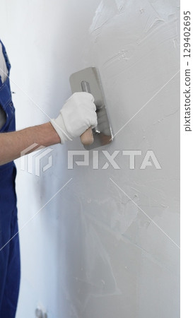 Professional construction worker wearing blue work overall is using a finishing trowel to carefully apply plaster on a wall, demonstrating expertise in home renovation and interior finishing 129402695