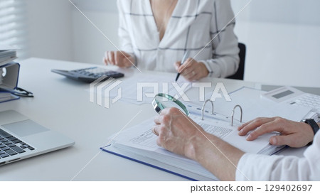 Two professional accountants working together, using a magnifying glass and a calculators, to audit financial reports in their brightly lit office. Audit and taxes in business 129402697