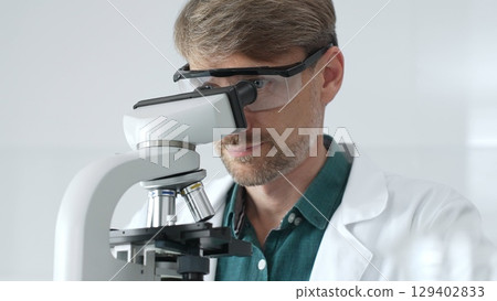 Senior man scientist wearing protective glasses and lab coat analyzing samples with a microscope in laboratory setting. Medicine, health care and science concept 129402833