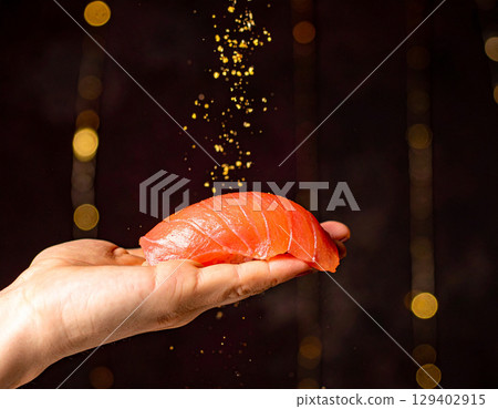 AI generated close-up of a large salmon sushi piece placed on back of a cupped hand with shimmering gold dust falling from above in an elegant dimly-lit dining room 129402915