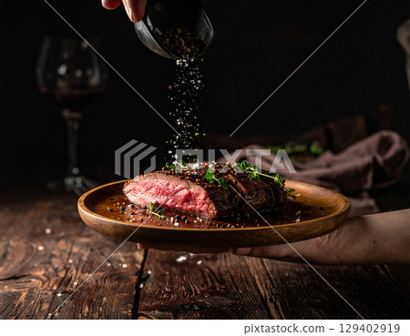 AI generated close-up of a perfectly cooked medium rare steak resting on a hand-held plate, garnished with falling black pepper and salt in an intimate dimly-lit dining setting 129402919
