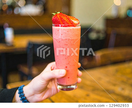AI generated refreshing strawberry smoothie in a glass held by hand with fresh strawberry pieces cascading down against a dimly-lit cozy cafe background 129402933