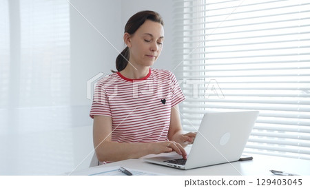 Focused female freelancer working from home, using laptop and sitting at white desk near window with blinds, concentrating on her online work. Business people concept Focused female freelancer working from home, using laptop and sitting at white desk near window with blinds, concentrating on her online work. Business people concept 129403045