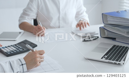 Two accountants collaborating in a well lit office, analyzing financial data and preparing reports with calculators and pens. Audit and taxes in business Two accountants collaborating in a well lit office, analyzing financial data and preparing reports with calculators and pens. Audit and taxes in business 129403125