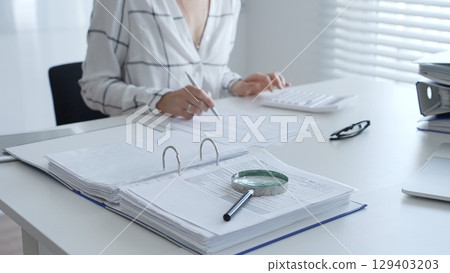 Female accountant is working with financial documents, using calculator and taking notes, with a magnifying glass and a folder on a white table in a bright office. Audit and taxes in business 129403203