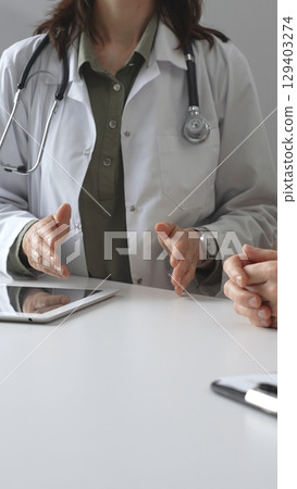 Doctor woman in medical coat is explaining diagnosis to patient man during a medical consultation in clinic office, gesturing with hands over a desk, vertical close up view. Medicine and health care 129403274