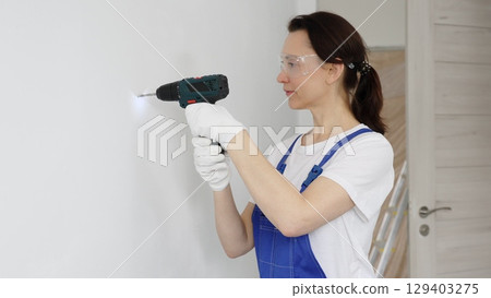 Woman worker wearing blue overall, safety glasses and gloves, is using cordless drill to make hole in white wall during renovation work. Horizontal portrait view 129403275