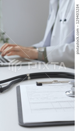 Stethoscope and medical records are on the desk opposite the doctor is typing on a laptop with a in a medical office, suggesting online healthcare or telemedicine services. Medicine concept 129403284