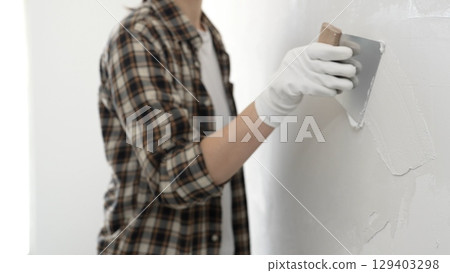 Unknown female construction worker wearing protective gloves and checkered shirt is spreading plaster on white wall during renovation project, smoothing surface with putty knife Unknown female construction worker wearing protective gloves and checkered shirt is spreading plaster on white wall during renovation project, smoothing surface with putty knife 129403298
