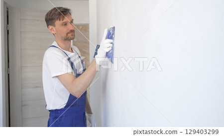 Man construction worker wearing protective white gloves and blue construction coveralls, is sanding white wall with blue hand block, preparing smooth surface during interior home renovation project 129403299